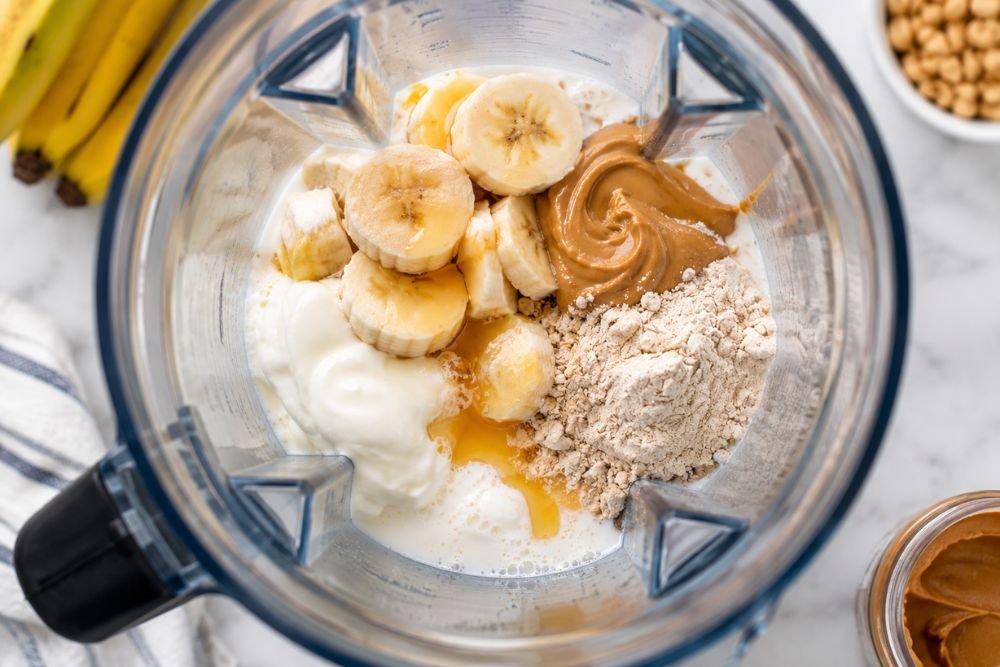 Smoothie ingredients including banana, peanut butter, Greek yogurt, milk, and protein powder inside a blender.
